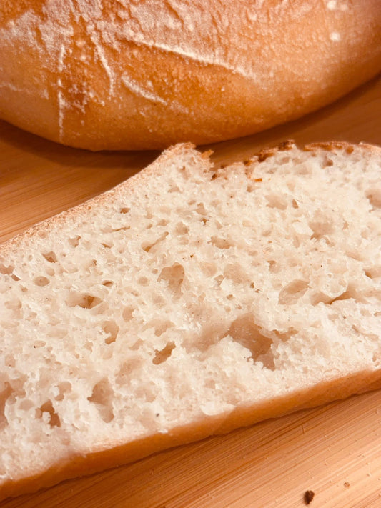 A close-up of a slice of Gluten Free bread with a focus on its porous texture, with a loaf partially visible in the background.